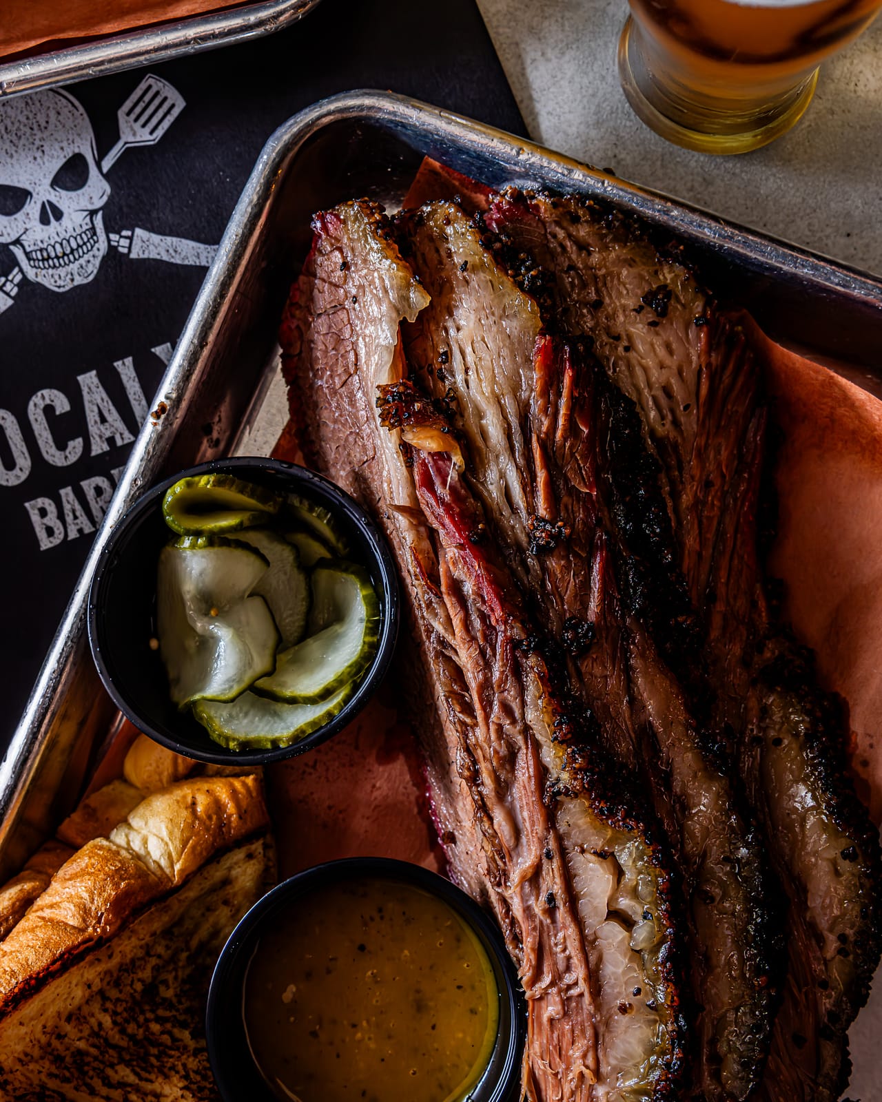Sliced Demkota prime brisket on a skull-branded tray with house pickles and mojo mustard at Apocalypse BBQ Kendall